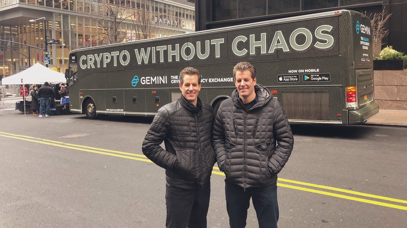 The founders of Gemini, Winklevoss twins, standing in front of a bus with a Gemini ad on it.